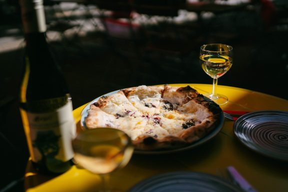 Pizza Capra mit zwei Gläsern Weißwein in der Abendsonne auf einem gelben Terrassentisch